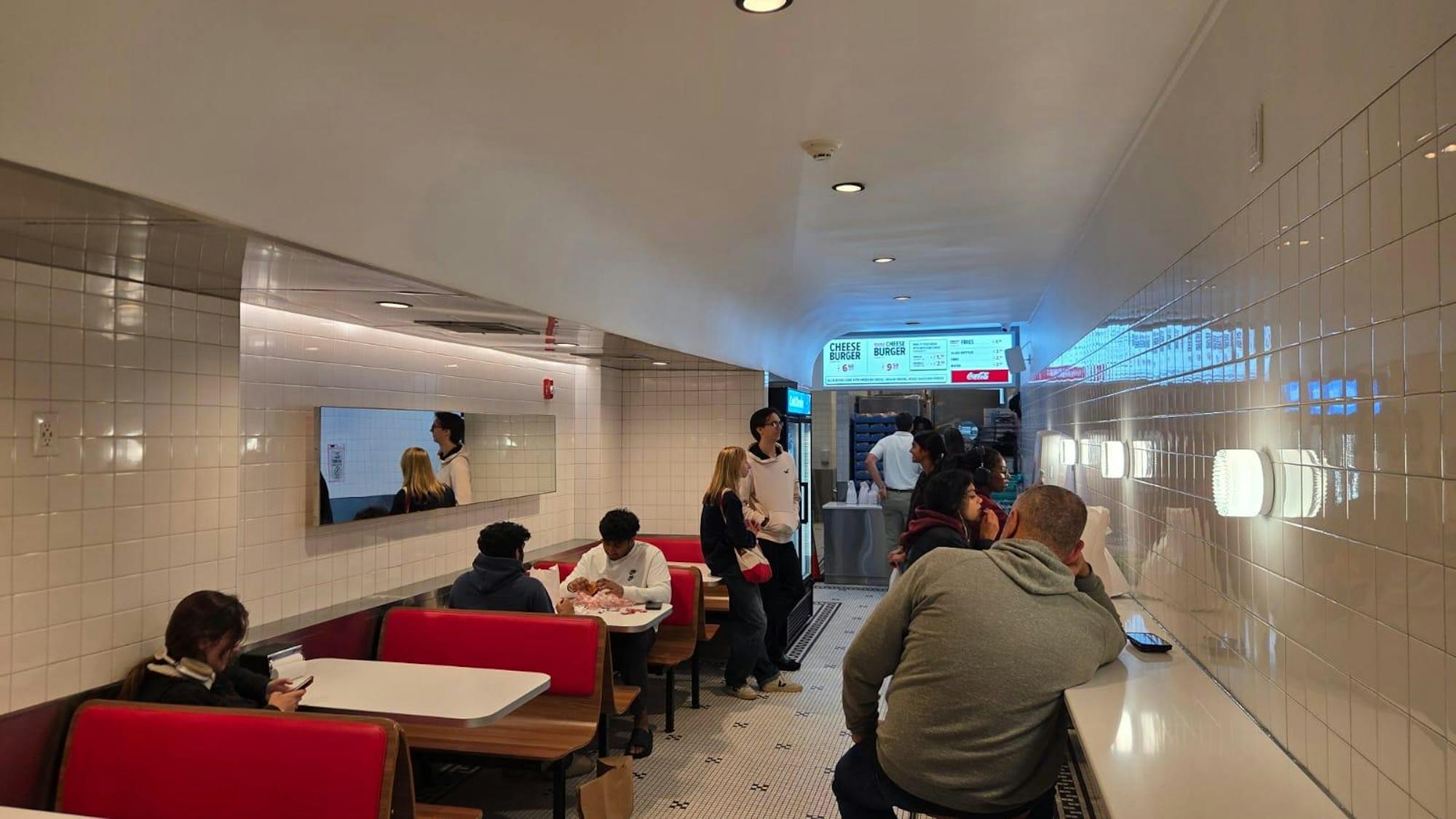 The interior of a narrow room with tiled white walls. There are multiple booths lining one wall with red cushions. There is a white bar with stools is on the other side of the room. There are people scattered throughout the room. There is a counter to order at the far side of the room. There is an illuminated menu.