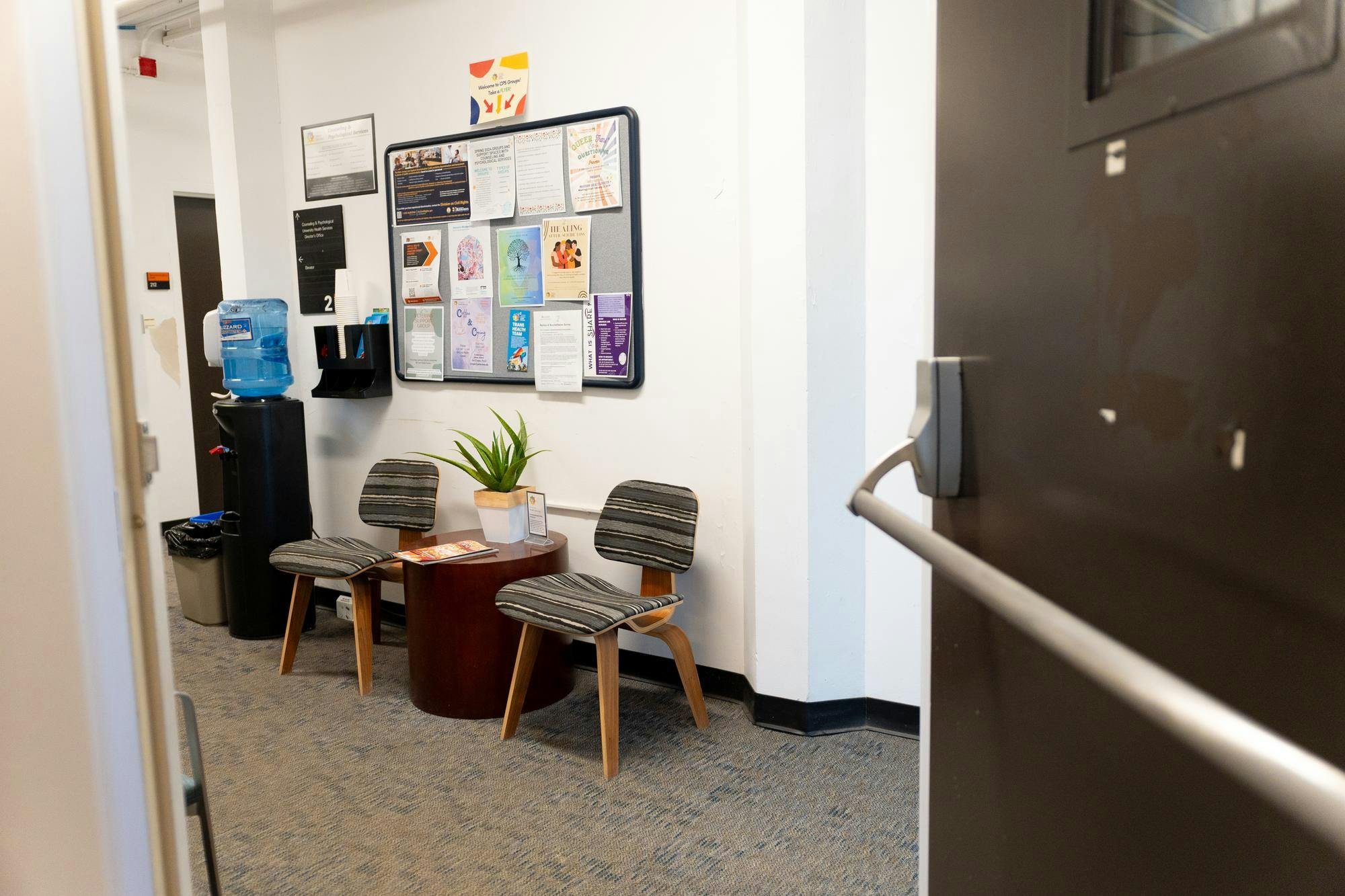 A waiting room with two chairs and a round table in the middle and a water cooler to the right. 