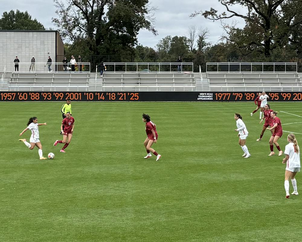 A Princeton player in a white jersey kicks a ball with Harvard defenders in red standing in front of her.