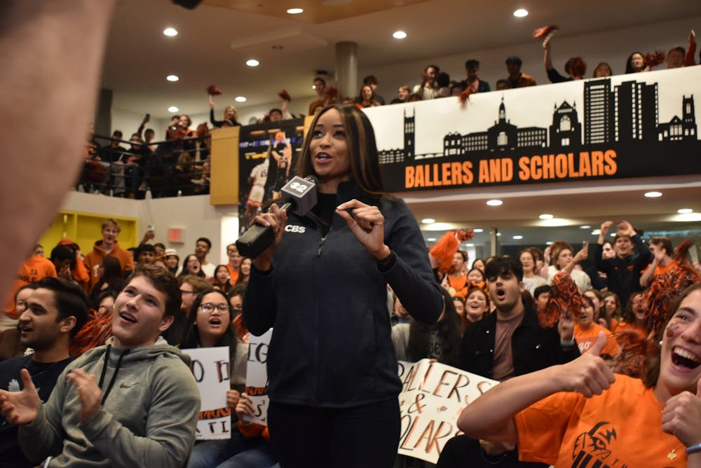 A woman wearing a CBS jacket speaks into a microphone in front of a crowd of cheering students dressed in orange