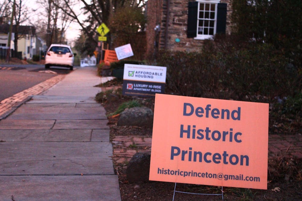 A rectangular orange sign reading "Defend Historic Princeton" in black lettering is planted on the lawn outside a home. Behind it, another sign, reading "Affordable Housing" with a green check mark, and "Luxury Hi-Rise Apartment Buildings Apartment BLDGS" with a red ex mark, is visible. 