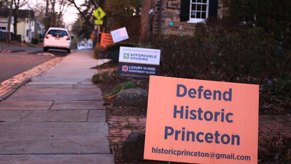 A rectangular orange sign reading "Defend Historic Princeton" in black lettering is planted on the lawn outside a home. Behind it, another sign, reading "Affordable Housing" with a green check mark, and "Luxury Hi-Rise Apartment Buildings Apartment BLDGS" with a red ex mark, is visible.