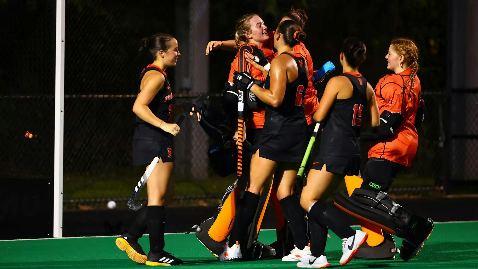 Field hockey players dressed in black uniforms with orange trim surround player in bright orange goalie pads while standing on green turf field with the night sky behind them.