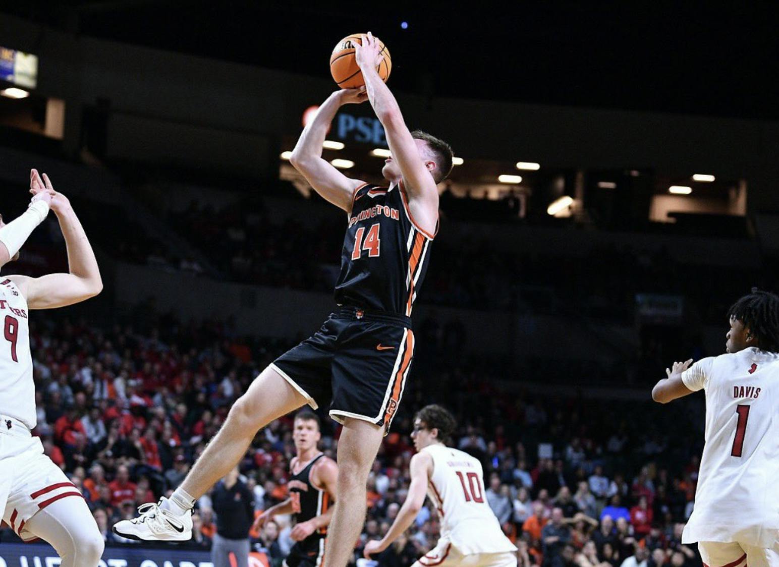 A male Princeton basketball player (14) with a ball in his hands ready to shoot surrounded by three men in white jerseys. 