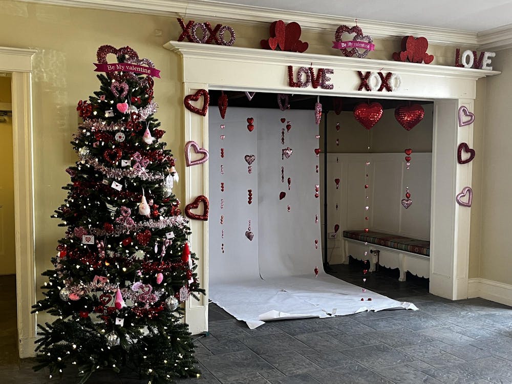 A white entry-way with red and pink heart decorations fixed to it. To its right, there is a green, artificial Christmas tree decorated with red, pink, and white Valentine's day decorations.