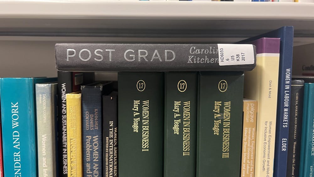 A black hardcover book with the title Post Grad, and the words Caroline Kitchener partially obscured by a library sticker, lying horizontally on top of a shelf of other books. 