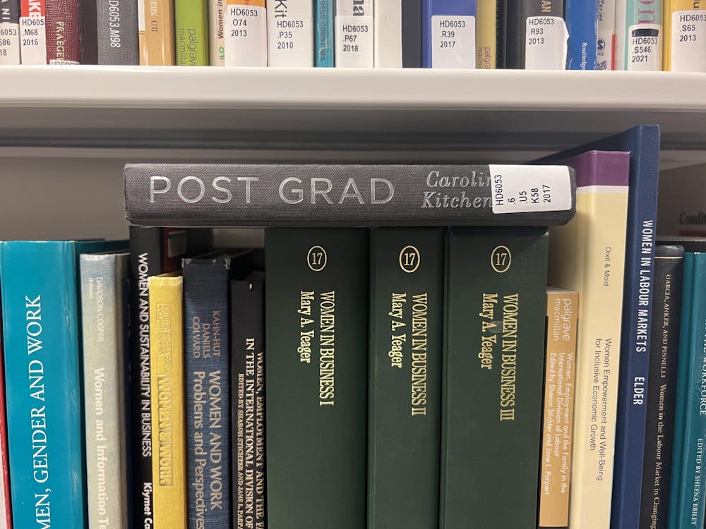 A black hardcover book with the title Post Grad, and the words Caroline Kitchener partially obscured by a library sticker, lying horizontally on top of a shelf of other books. 