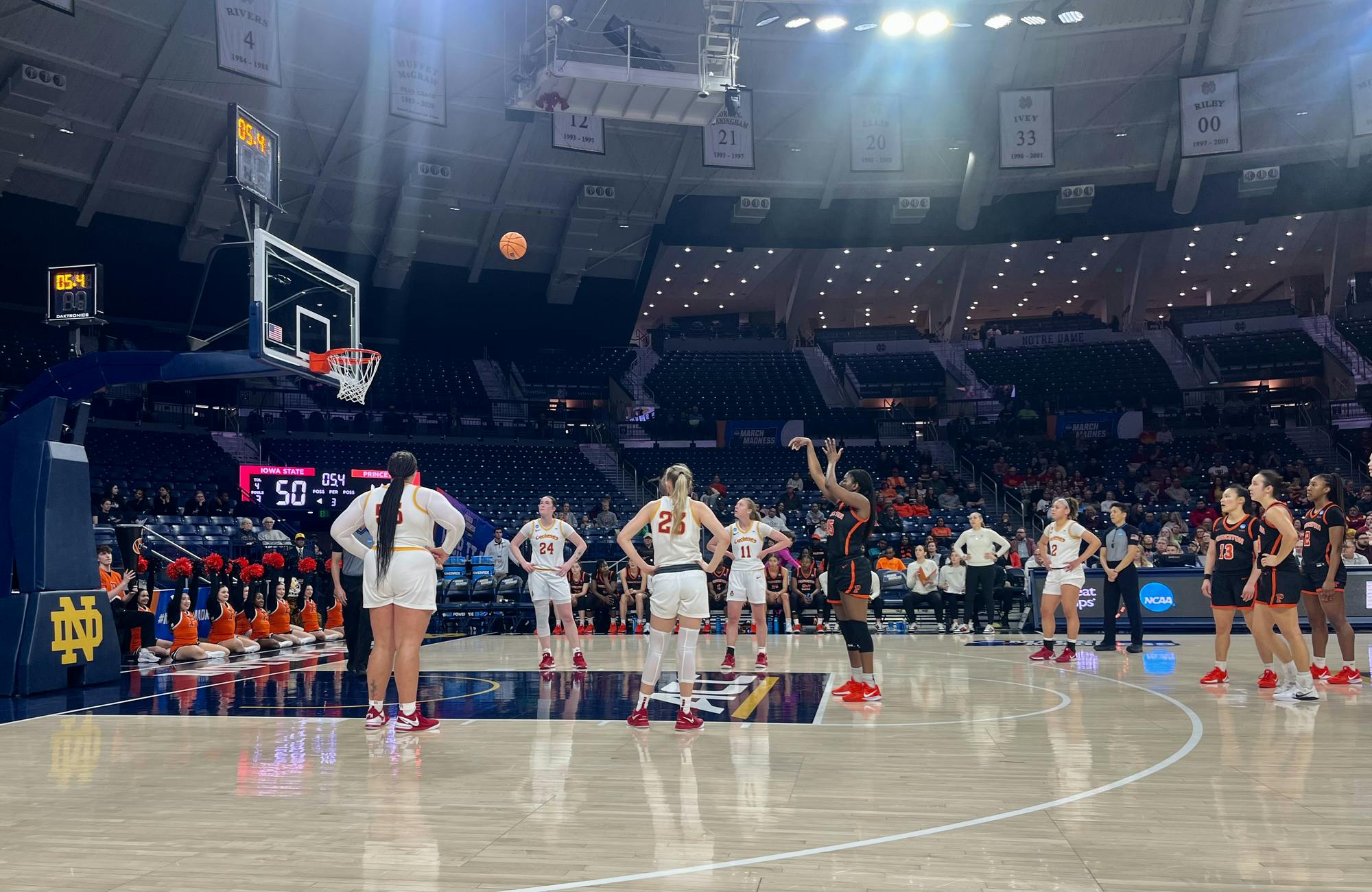 A woman wearing a basketball uniform shoots a basketball from the free-point line on a basketball court. Four other girls stand around. An arena is in the back. 