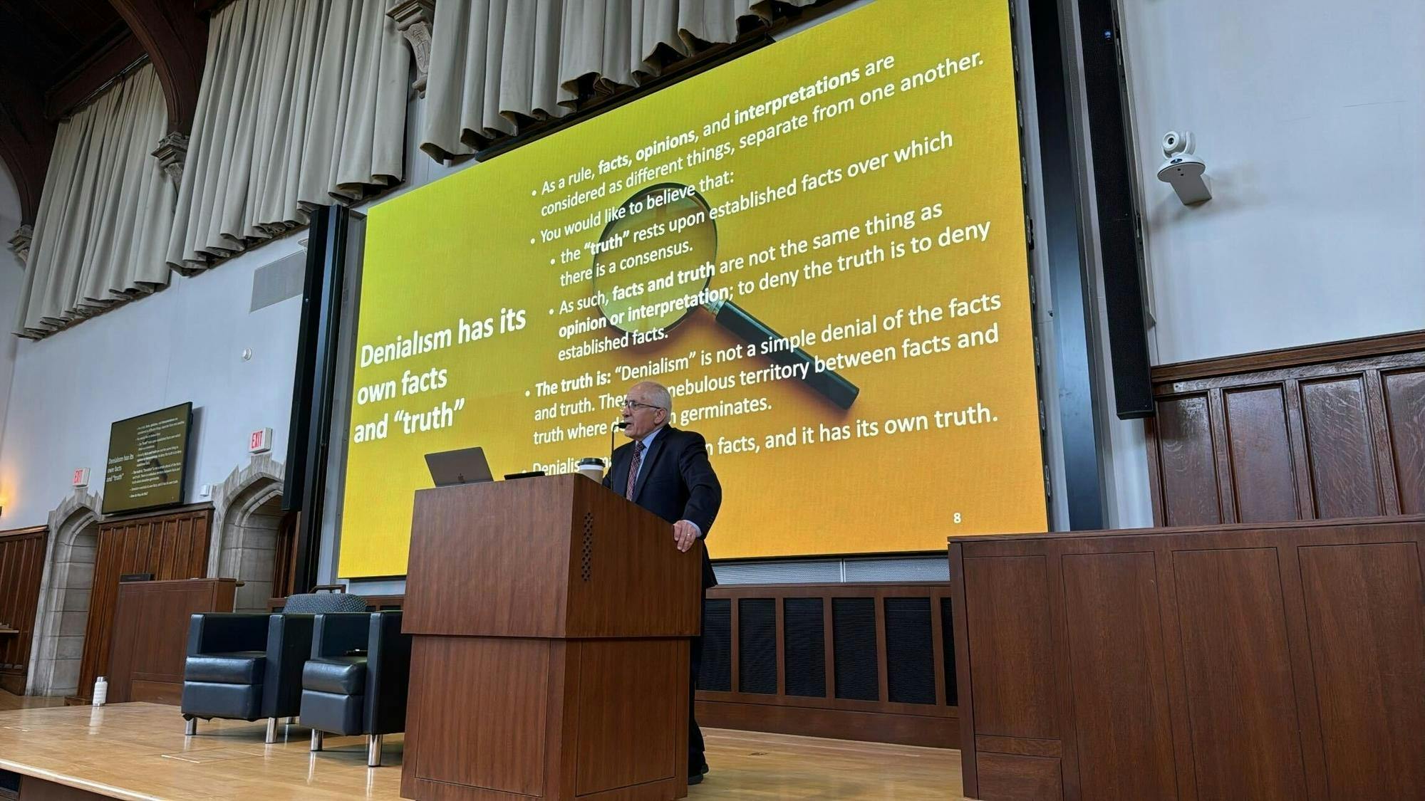 An elderly man gives a speech at a dais as a slideshow is displayed on the screen behind him. 