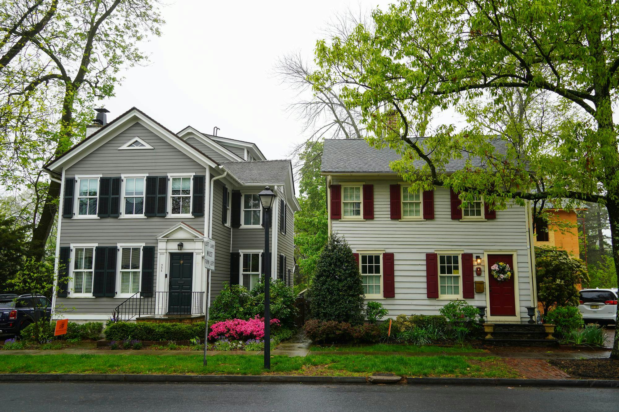 Two houses, one gray and one yellow, on a neighborhood street with green shrubbery lining the sidewalk.