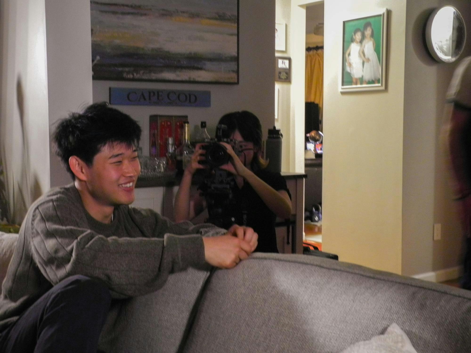 A person in a grey sweater is sitting on a grey couch. A person wearing a black skirt is holding a black camera.