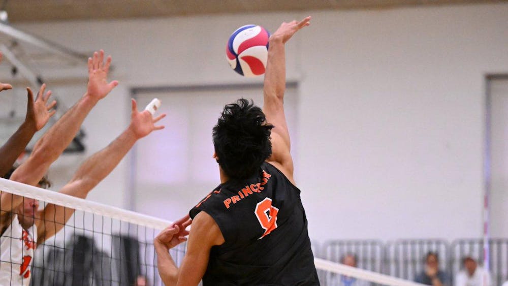A player in orange and black jumps up to spike volleyball, and two sets of blocking players meet him at the net. 