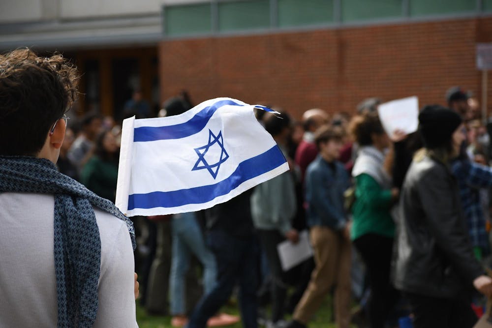 A person wearing a scarf and white shirt stands with their back to the camera holding the flag of Israel. In the background a group of students walk by.