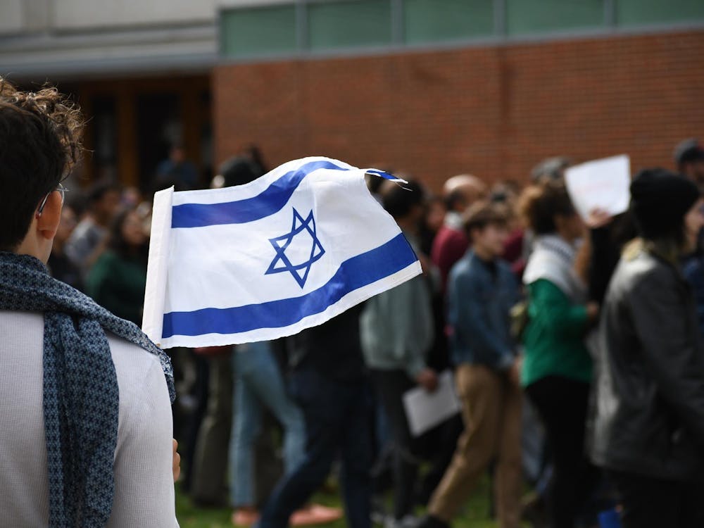A person wearing a scarf and white shirt stands with their back to the camera holding the flag of Israel. In the background a group of students walk by.