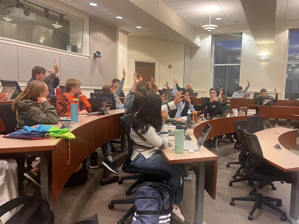 Students sit and raise their hands in black spinning desk chairs in front of three large wooden tables.