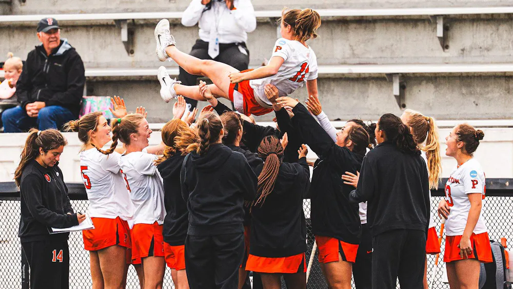 Lacrosse players in white and orange jerseys celebrating, lifting a player in the air.