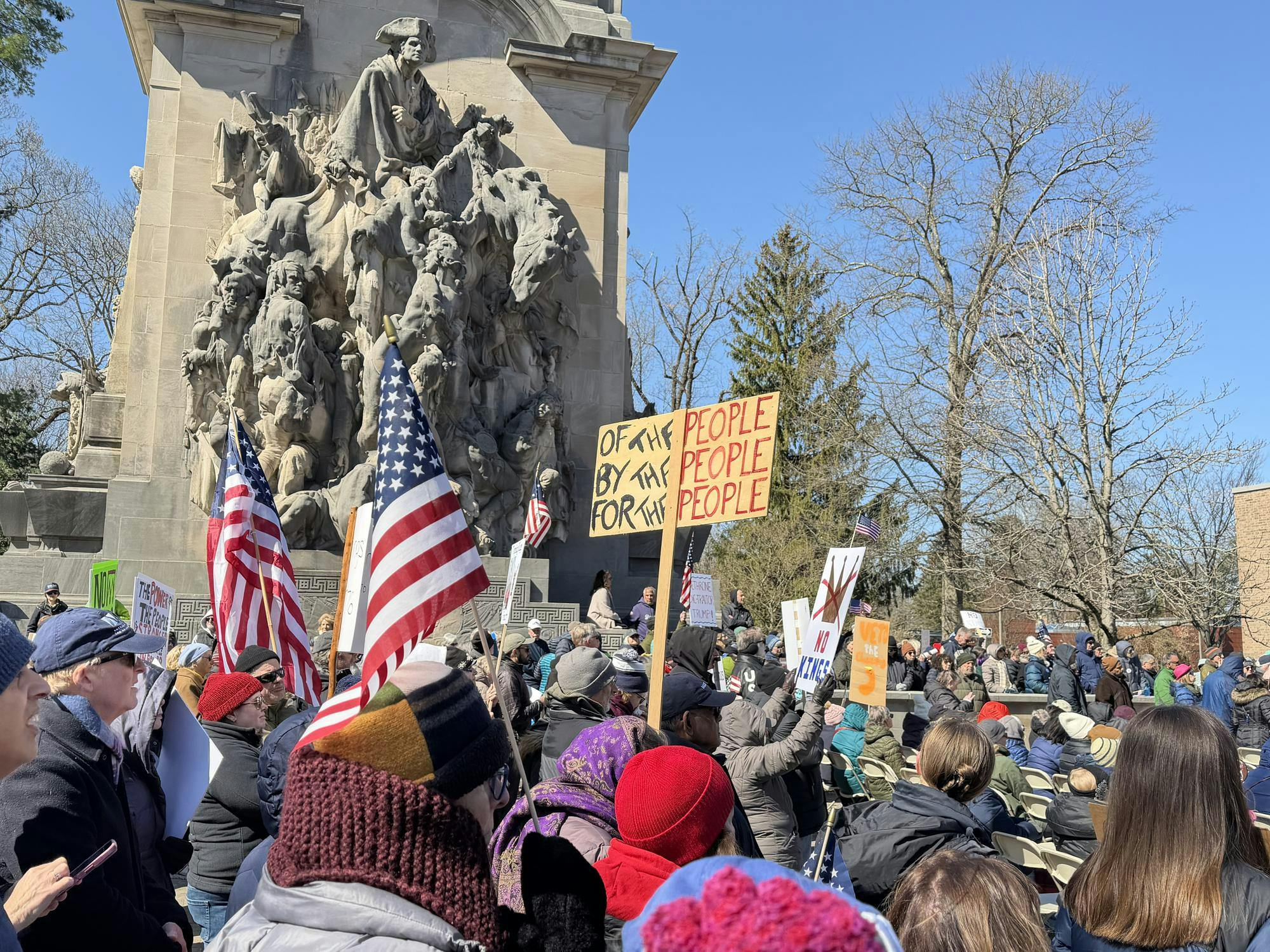 People with signs, standing around a battlefield statue.