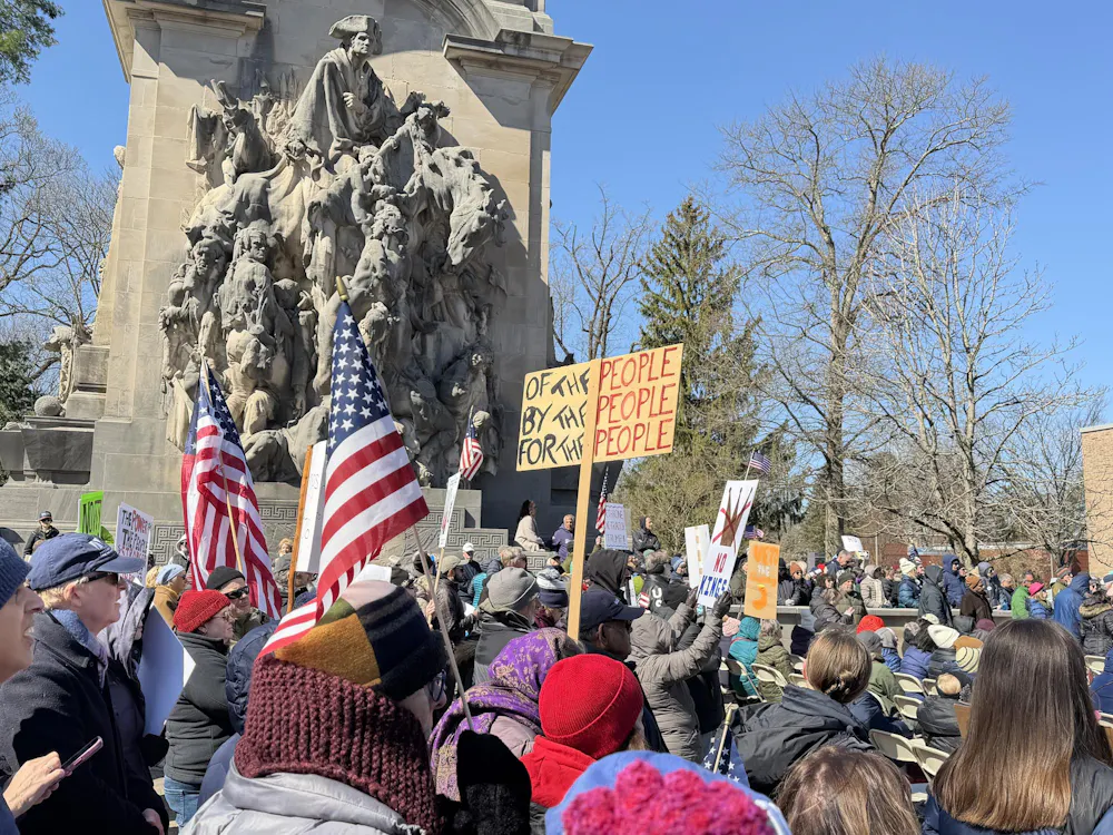 People with signs, standing around a battlefield statue.