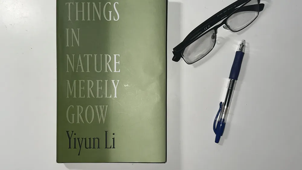 A green book with white letters that read "Things in Nature Merely Grow." There is a pair of black glasses and a black pen scattered next to the book. The items are all on a white background.