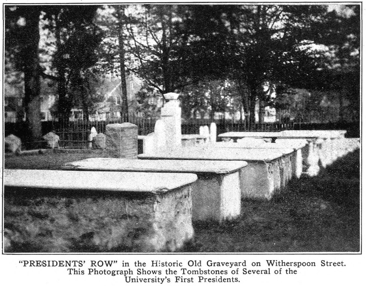 A black and white 1924 image of a row of mausoleums surrounded by other gravestones in a picketed cemetery