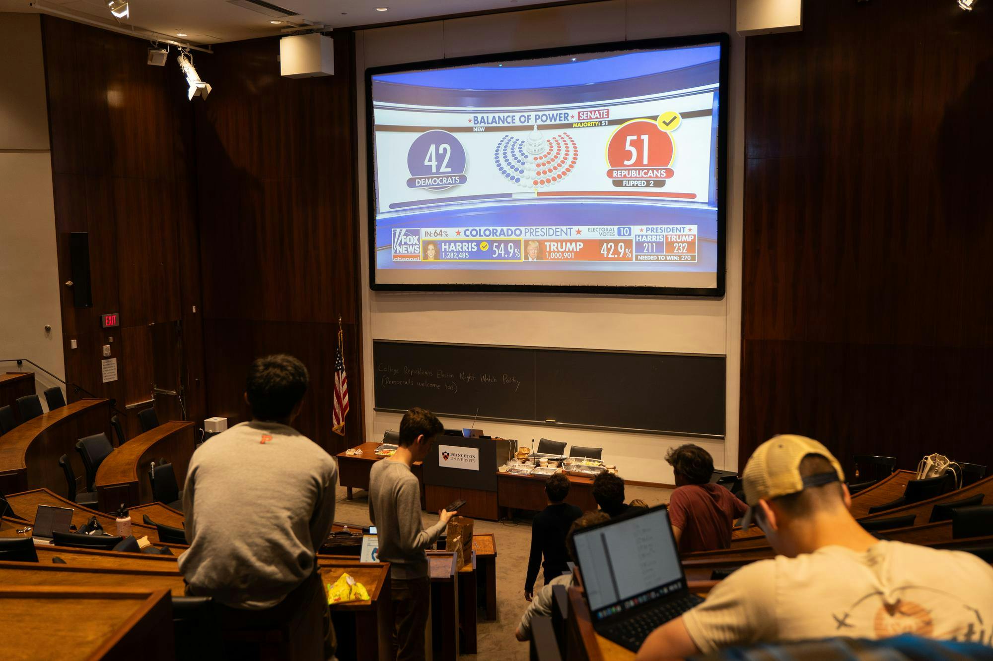 People gather in large lecture hall watching a large projection screen displaying Fox News.