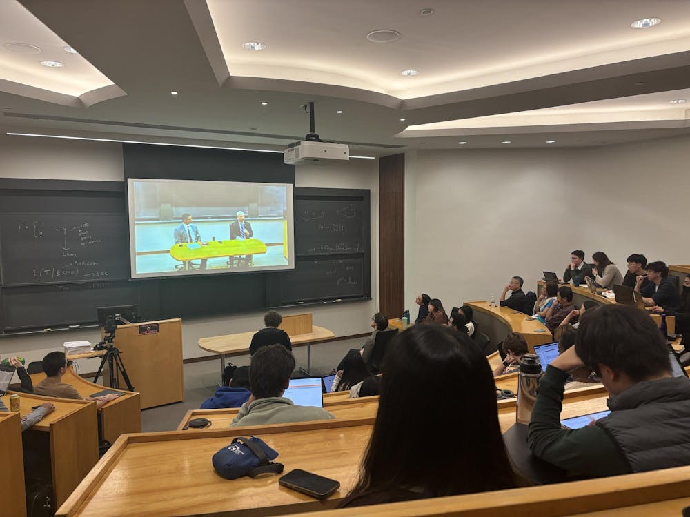 Student sit in a lecture hall and look at a screen featuring two men sitting and having a conversation. 