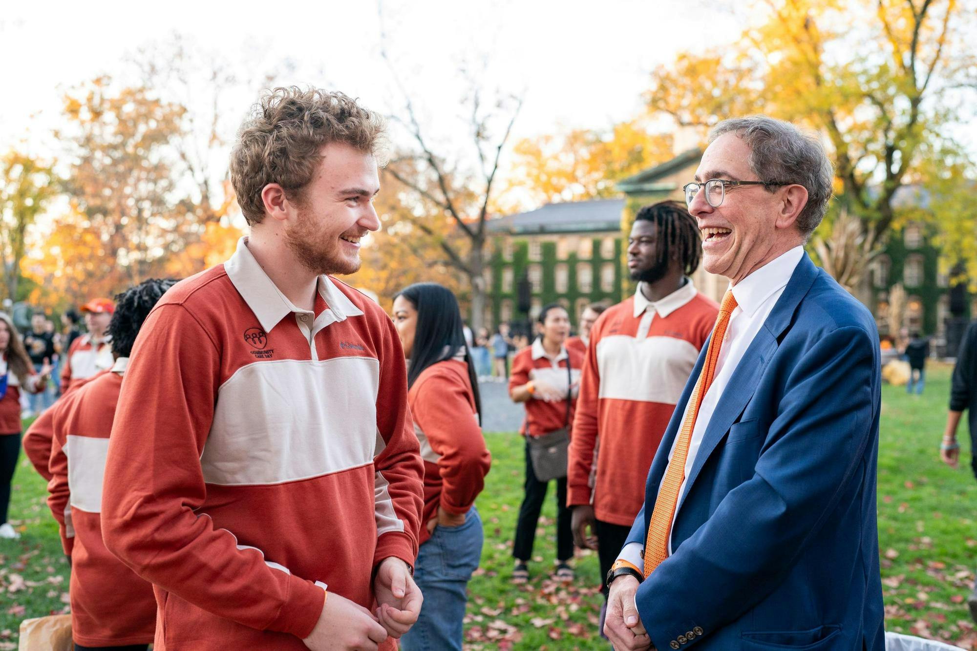 Two men smile and laugh on a grassy field in front of an old building. 