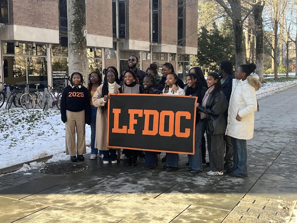 Fifteen students pose for a photo with an orange and black banner that reads “LFDOC” on a snowy stone walkway lined with trees.   