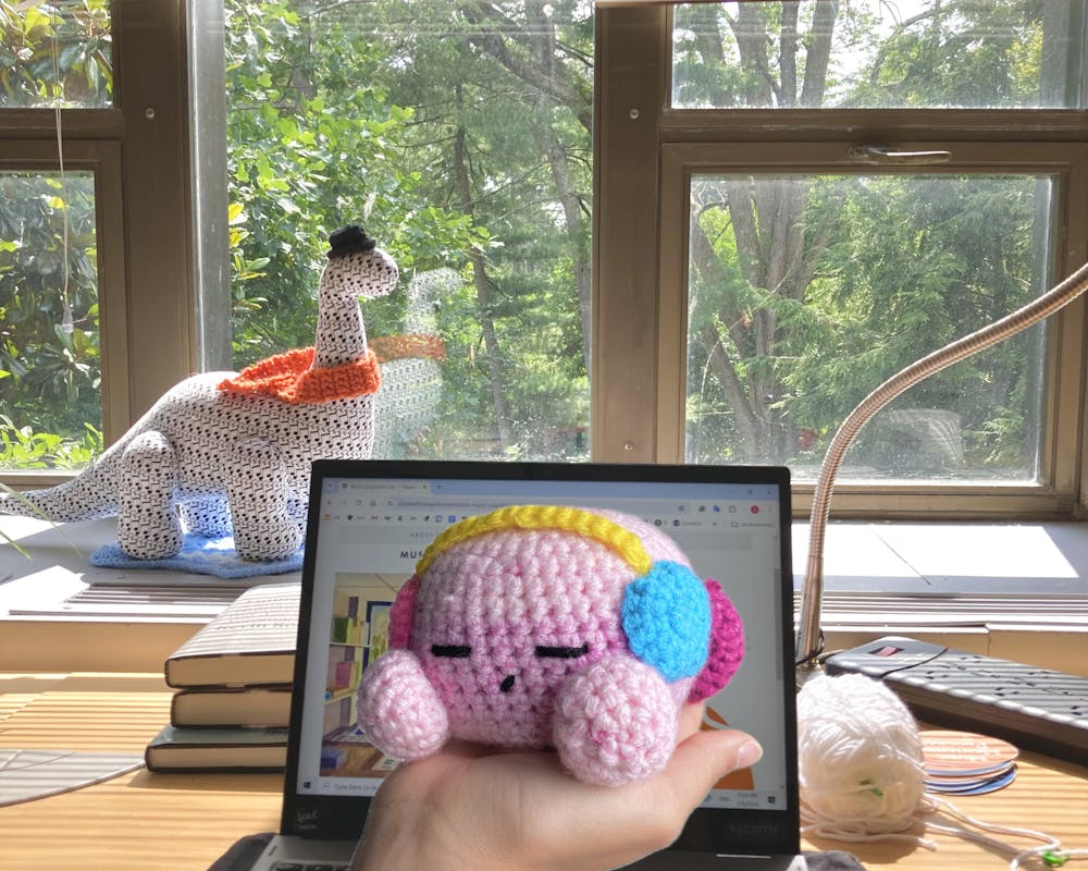 A crochet Kirby with headphones held in front of a window sill, upon which sits a crochet dinosaur, looking out into the foliage of trees. 