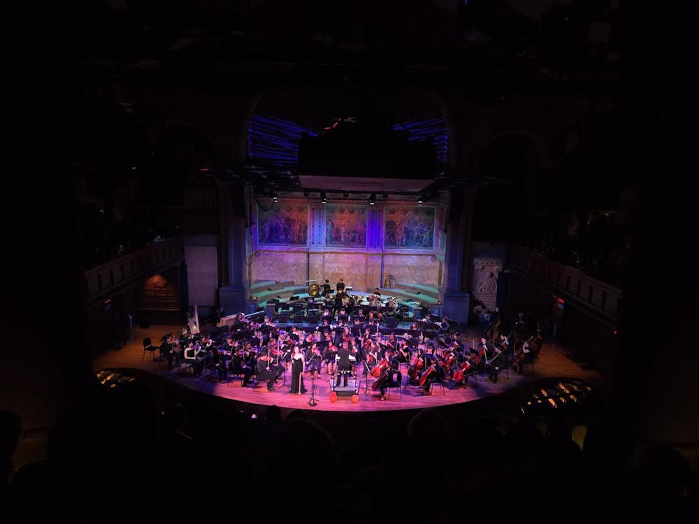 An orchestra gathered upon a large stage lit by pink and purple lights.  