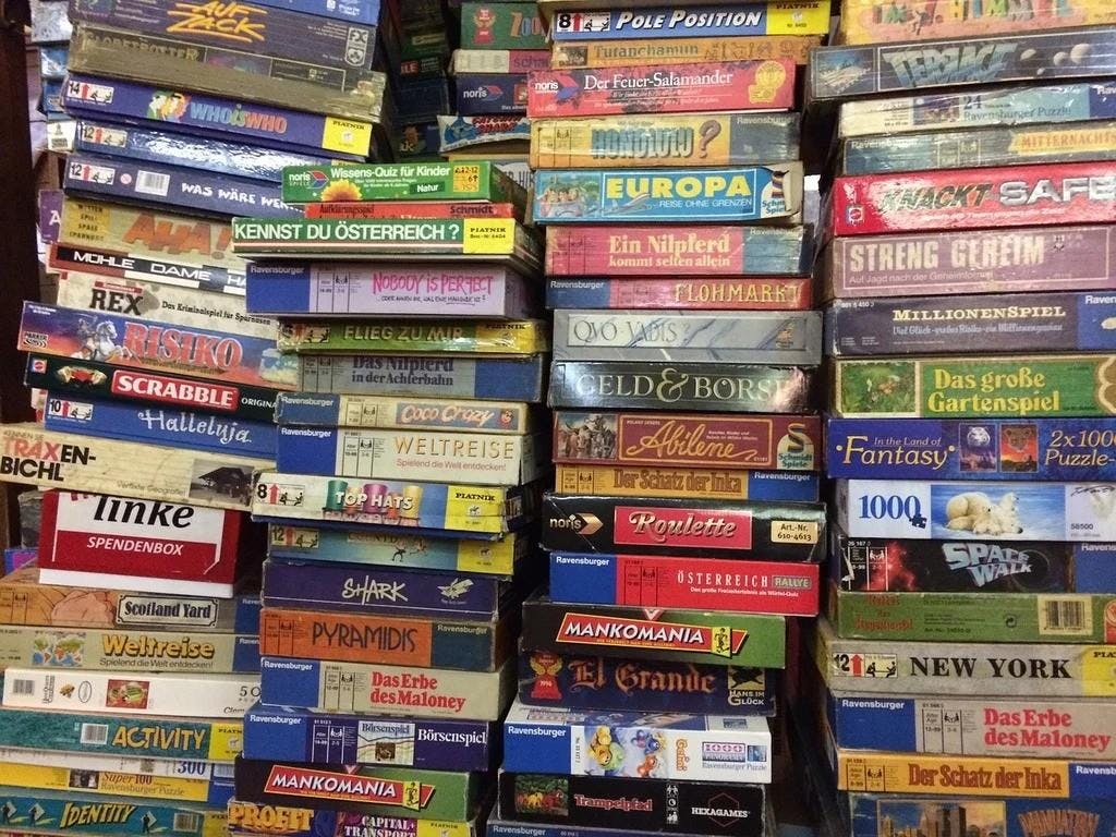 Four stacks of board games with about 15 in each stack with more in the background, many of them in German such as Ein Nilpferd and Weltreise.