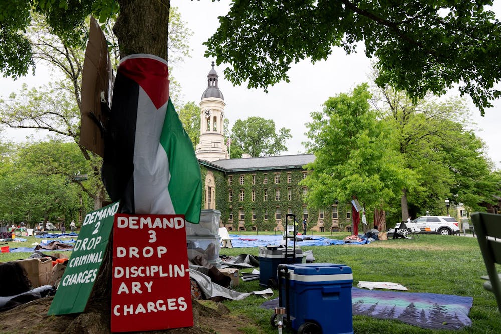 A black, red, white, and green flag wrapped around a tree with two signs at its base which read "demand 2 drop criminal charges" and "demand 3 drop disciplinary charges"