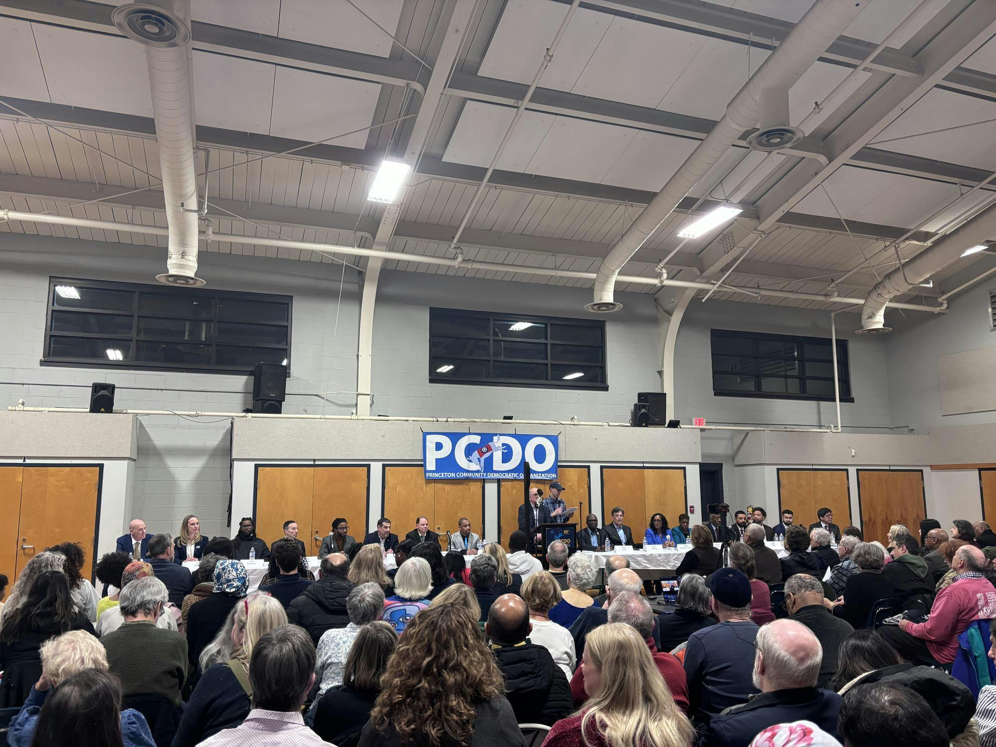 A crowd faces a stage with seated candidates. There is a podium in the middle of the stage, as well as a PCDO banner above it. 