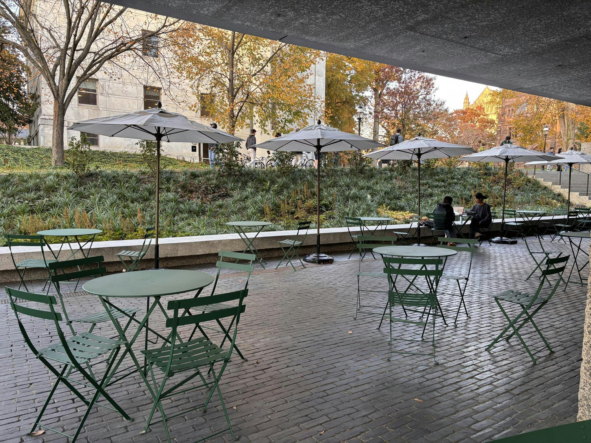 Metal green chairs and tables sit on a stone pavilion with autumn trees in the background. 