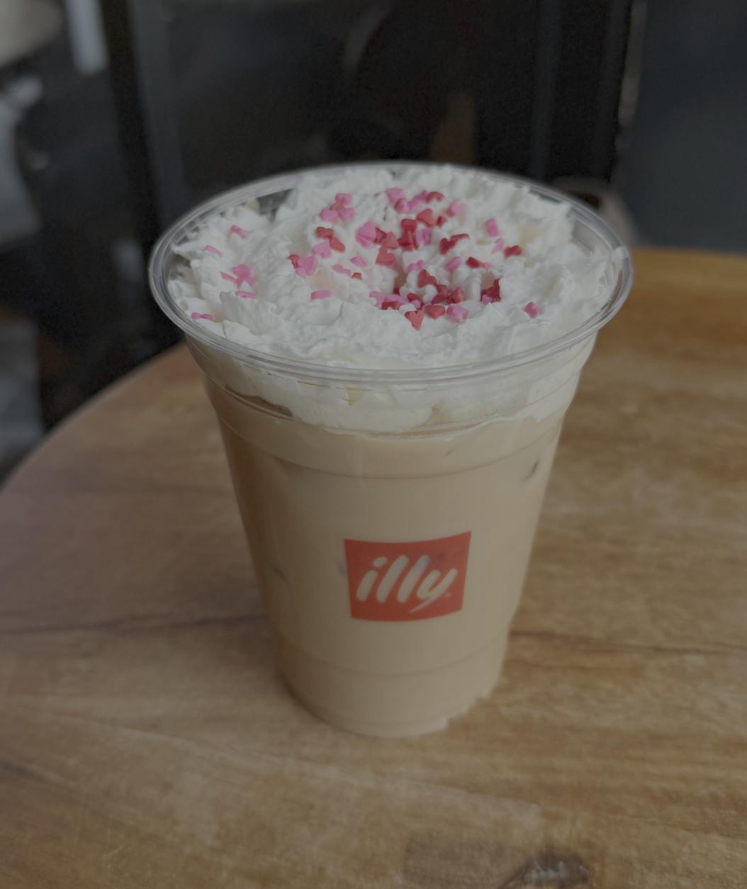 An iced latte sits on a wooden table, topped with whipped cream and heart-shaped pink and red sprinkles. 