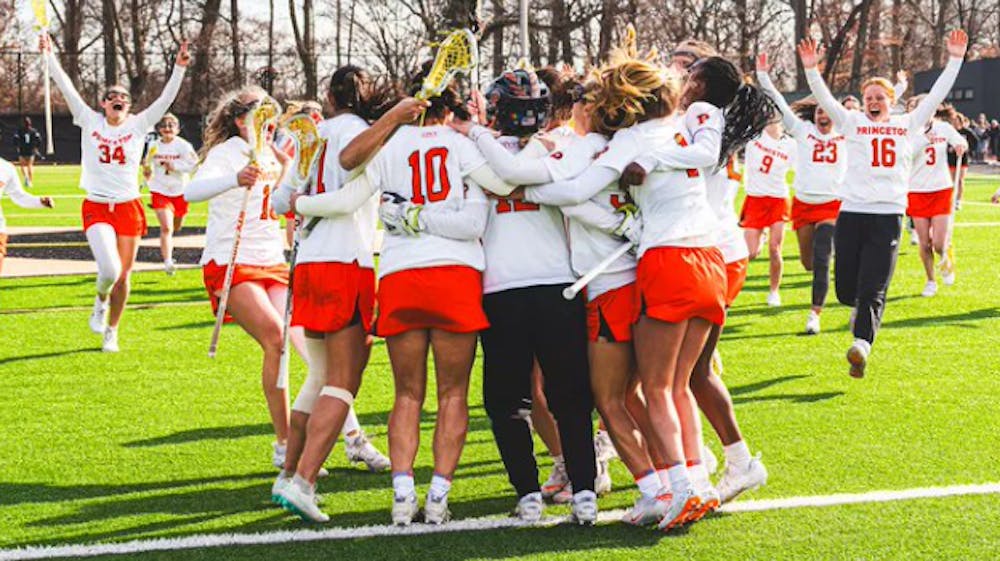 Princeton women's lacrosse in white jerseys celebrating in a huddle.