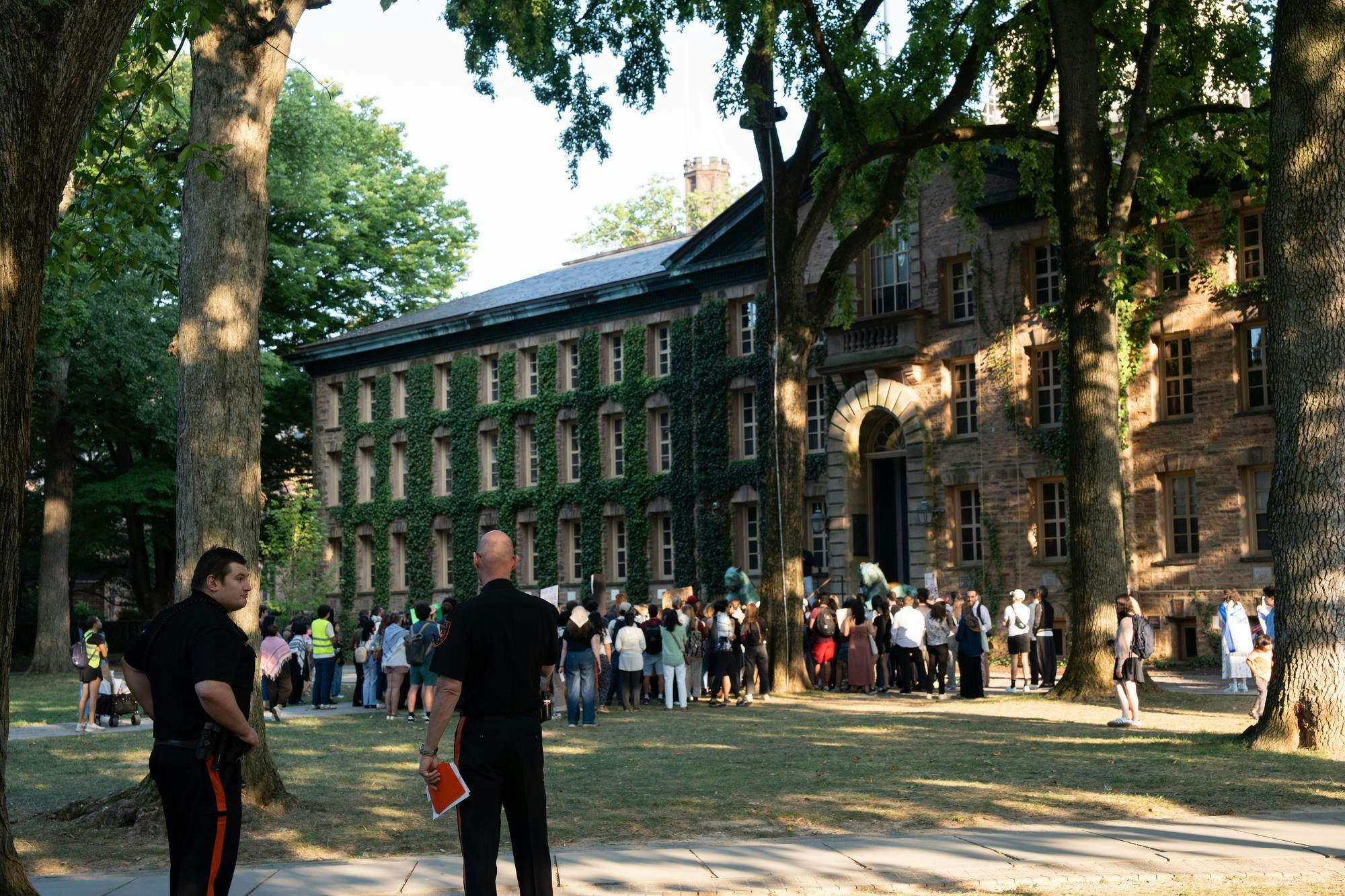In front of a large, ivy-colored building, a group of protesters are gathered. Some are holding signs. In the foreground, two Public Safety (PSAFE) officers are standing and talking to one another.