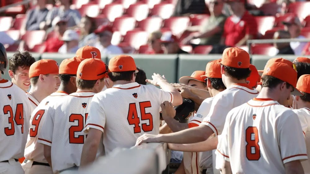 Princeton baseball players in white jerseys and orange baseball hats put their hands in a circle for a team chant.