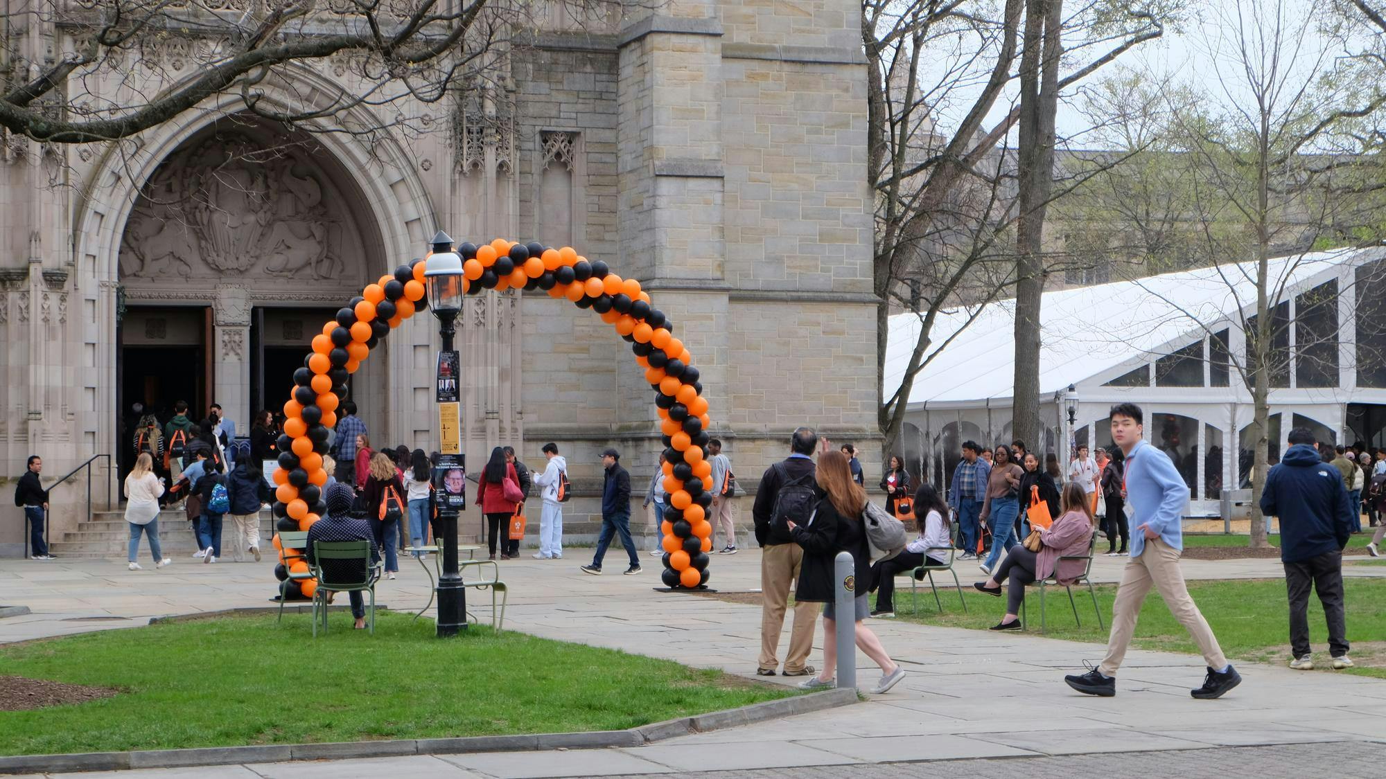 A large stone building is stood behind an orange and black balloon arch. A number of people are milling about the area.