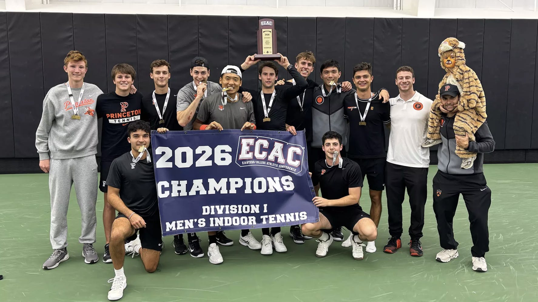 A group of Princeton tennis players holding a poster that says ‘champions,’ and a player in the center of the photo is hoisting a trophy