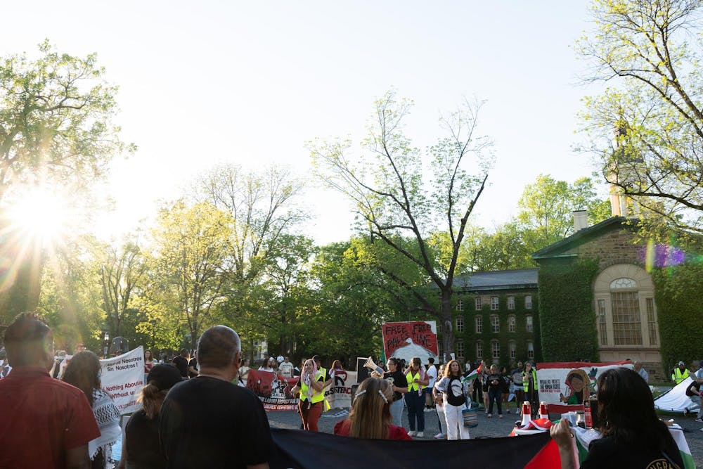 A group of protesters gather in a circle outside in front of a large building covered in ivy. Someone speaks into a megaphone, and some protesters carry signs.