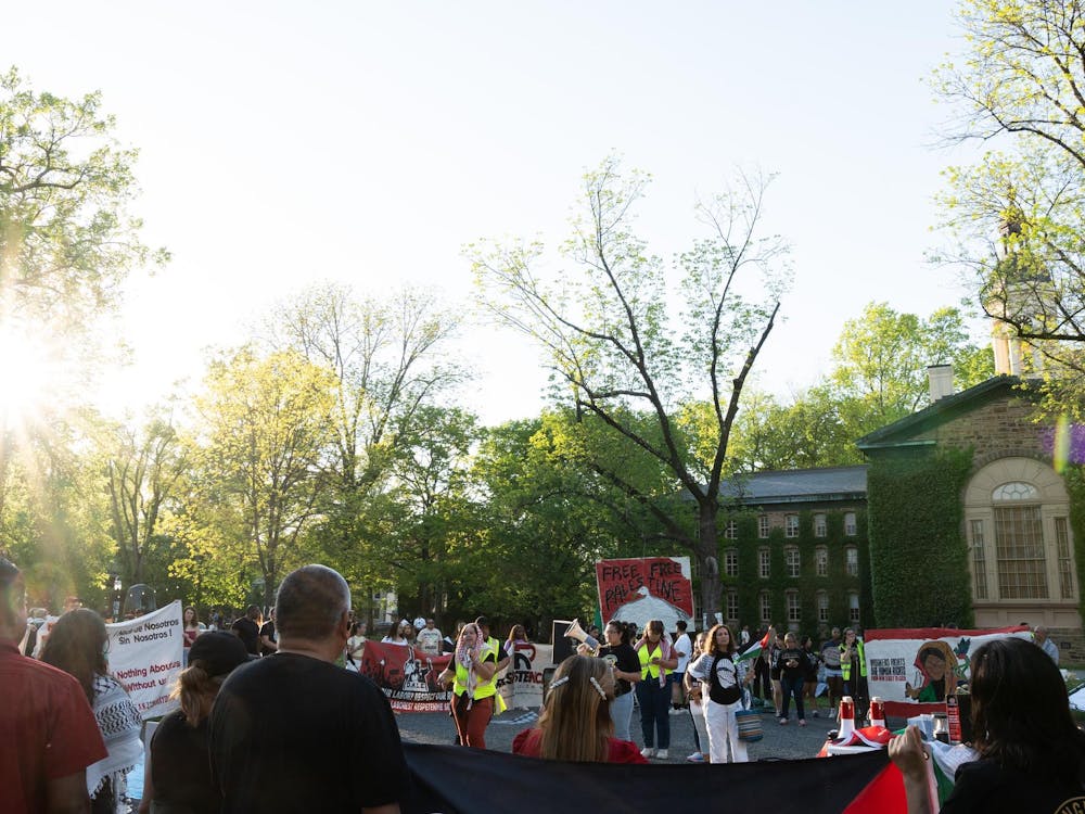A group of protesters gather in a circle outside in front of a large building covered in ivy. Someone speaks into a megaphone, and some protesters carry signs.