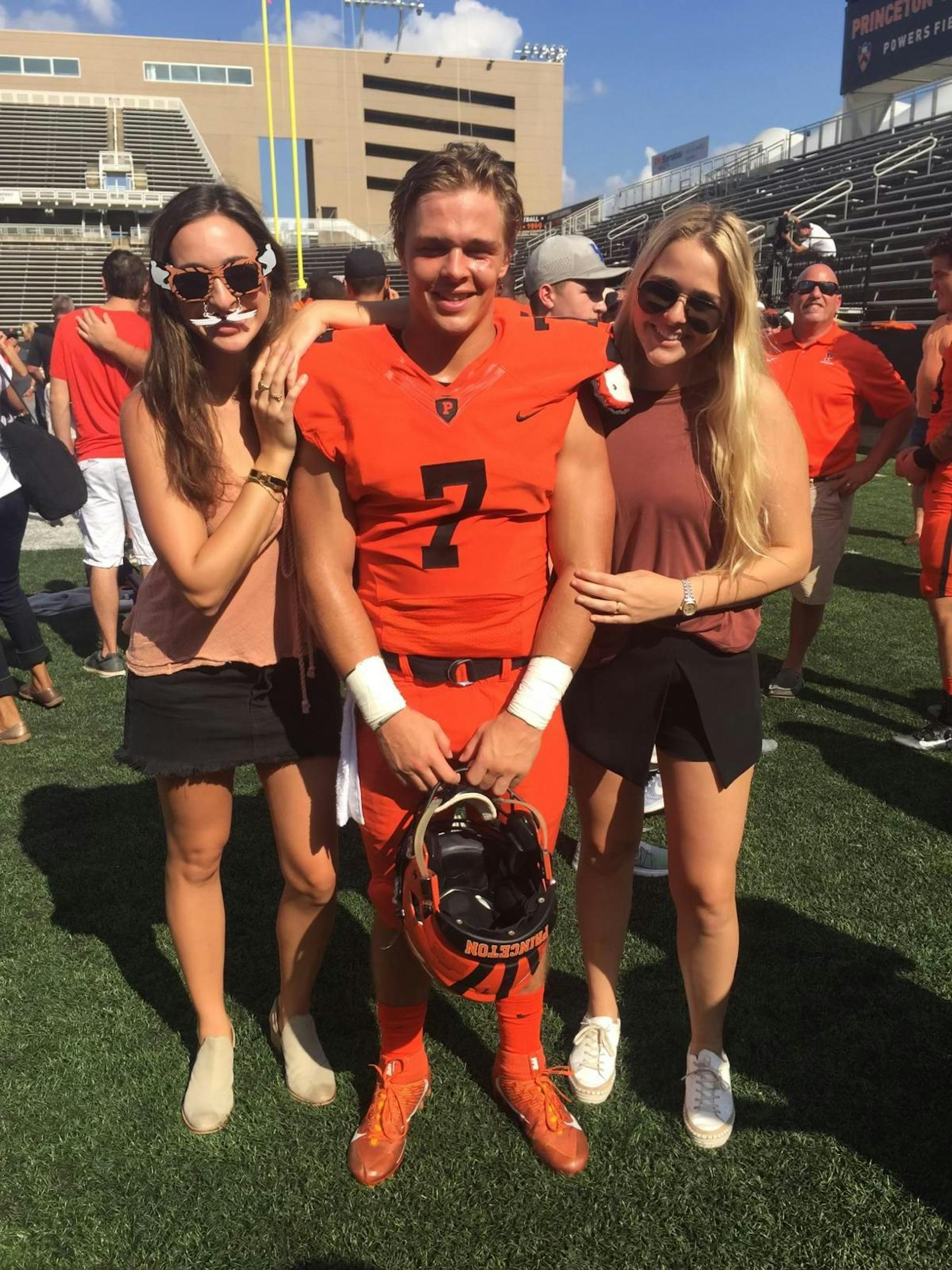 Ginnie, Tiger, and Sophie Bech stand in a line facing the camera. Tiger is wearing his football uniform, and in the background is the Princeton Stadium.