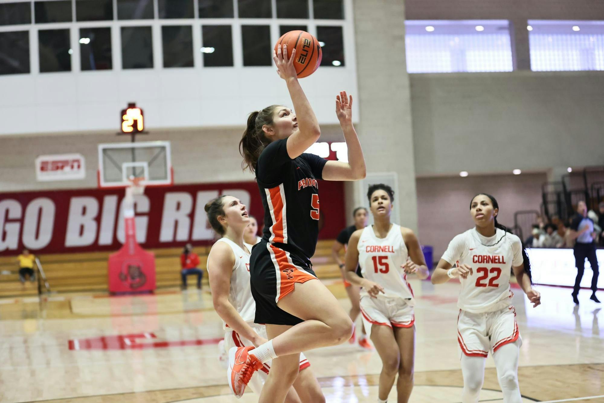 Player in orange and black jumps to attempt a layup with three defenders in white uniforms looking on. 