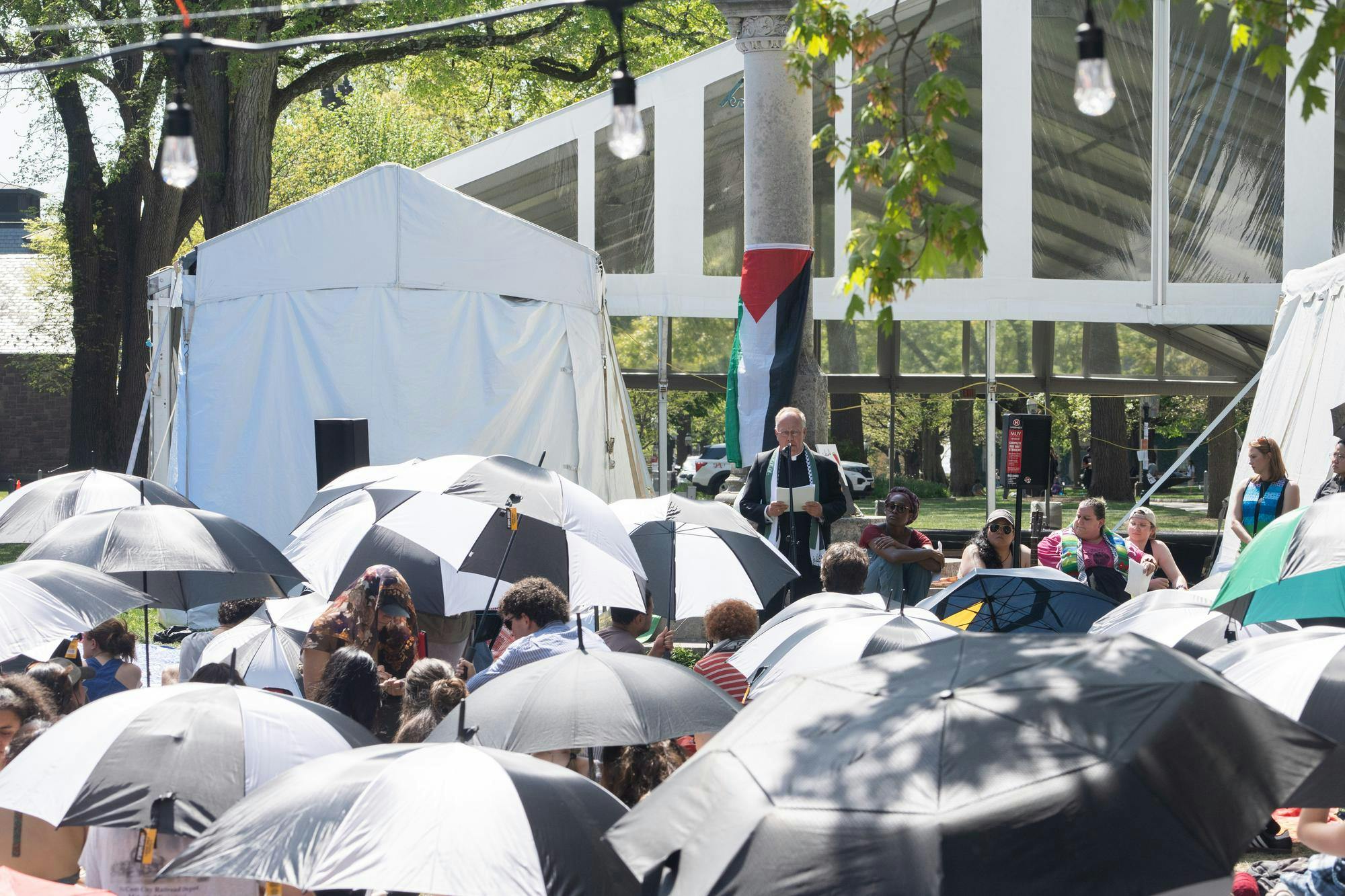 Several black and white umbrellas are at the sit-in on the McCosh courtyard. In the background, a speaker in black and white robes speaks in front of a pole draped in a green, red, and black Palestinian flag.