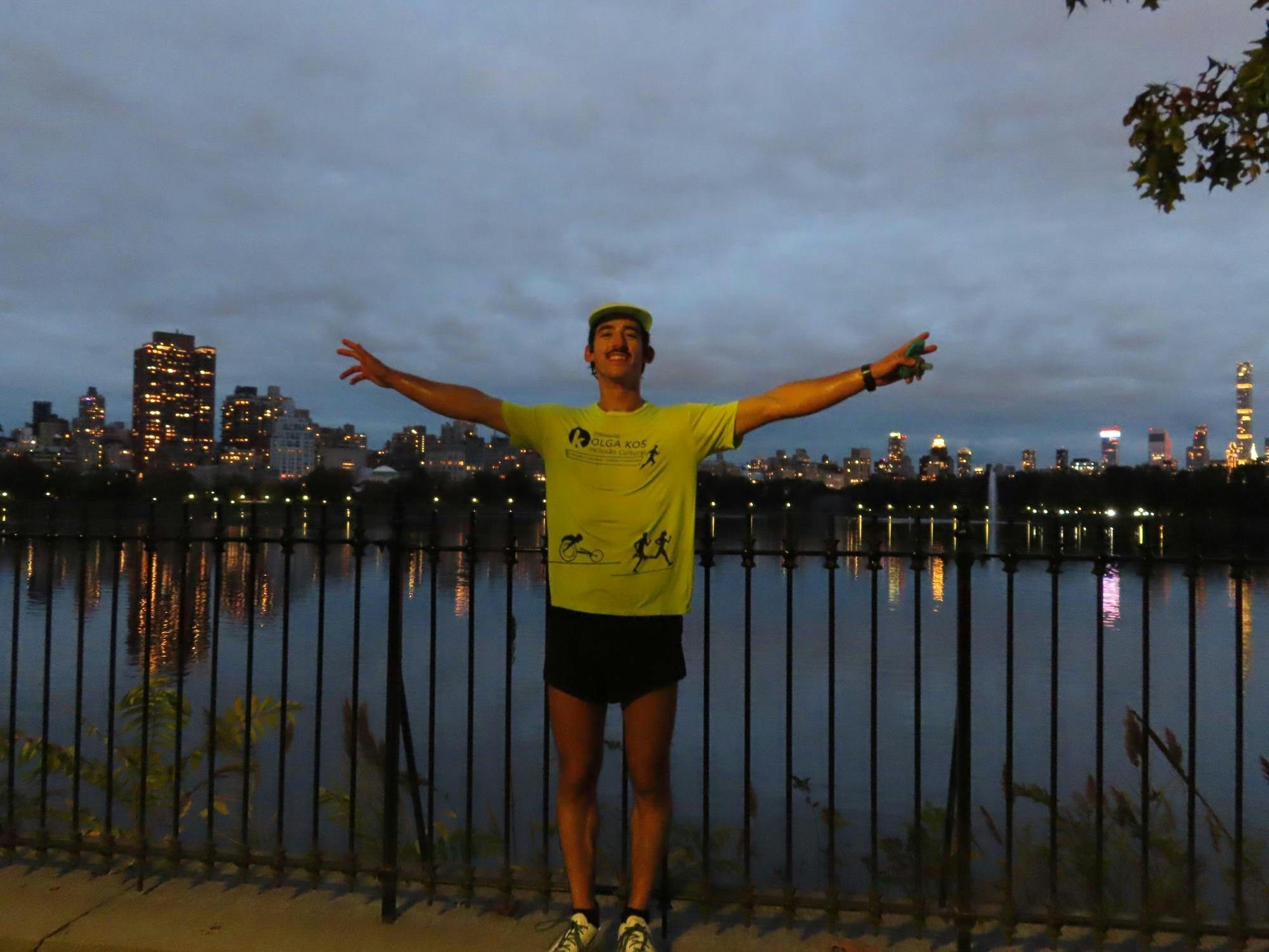 A man stands wearing a bright yellow t-shirt, yellow cap, and black shorts, arms spread wide, with a big smile on his face, in front of reservoir lined with buildings. 