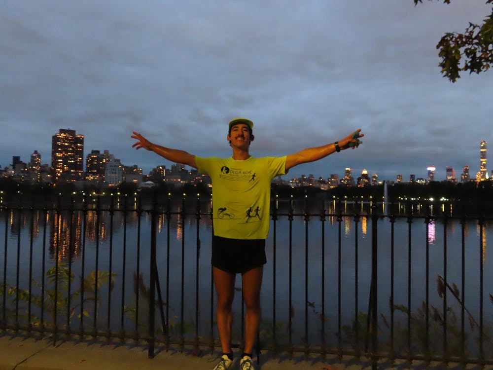 A man stands wearing a bright yellow t-shirt, yellow cap, and black shorts, arms spread wide, with a big smile on his face, in front of reservoir lined with buildings. 