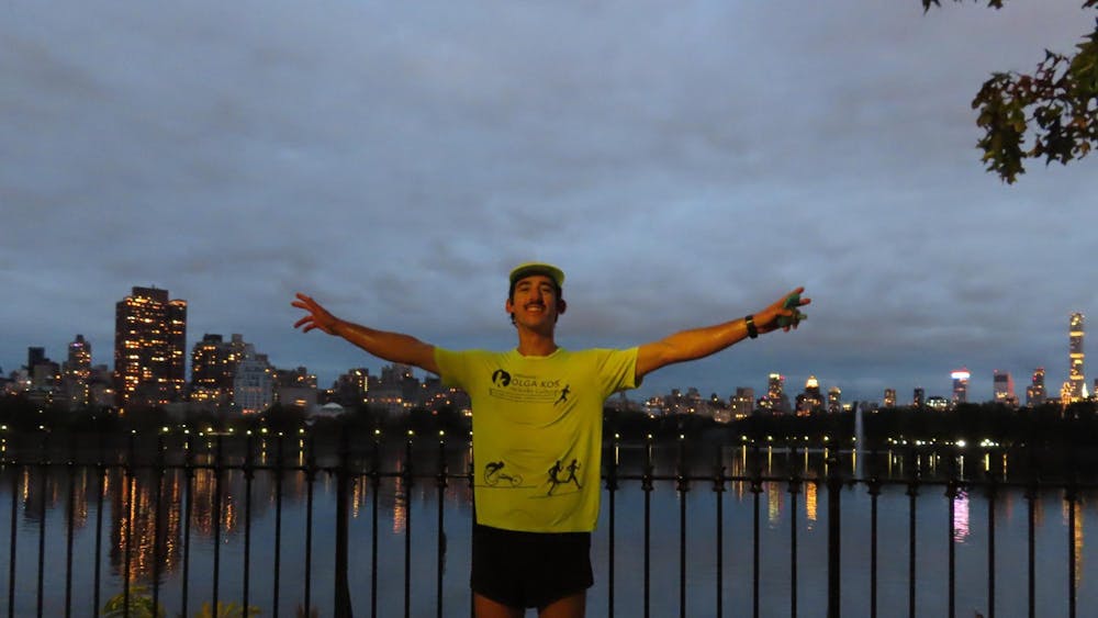A man stands wearing a bright yellow t-shirt, yellow cap, and black shorts, arms spread wide, with a big smile on his face, in front of reservoir lined with buildings.