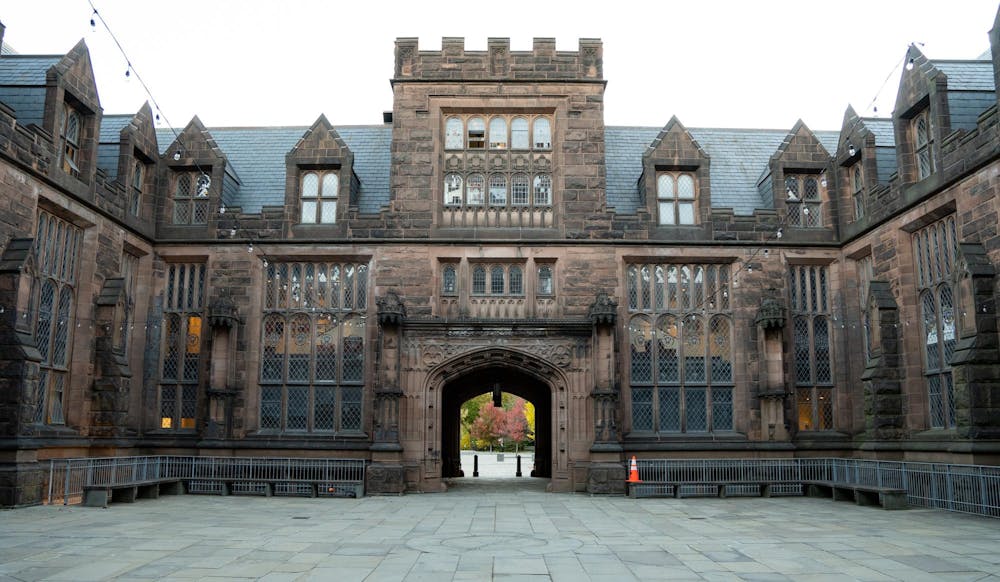 A building of brown stone around a courtyard. A tree is in the distance through an archway. It is fall. 