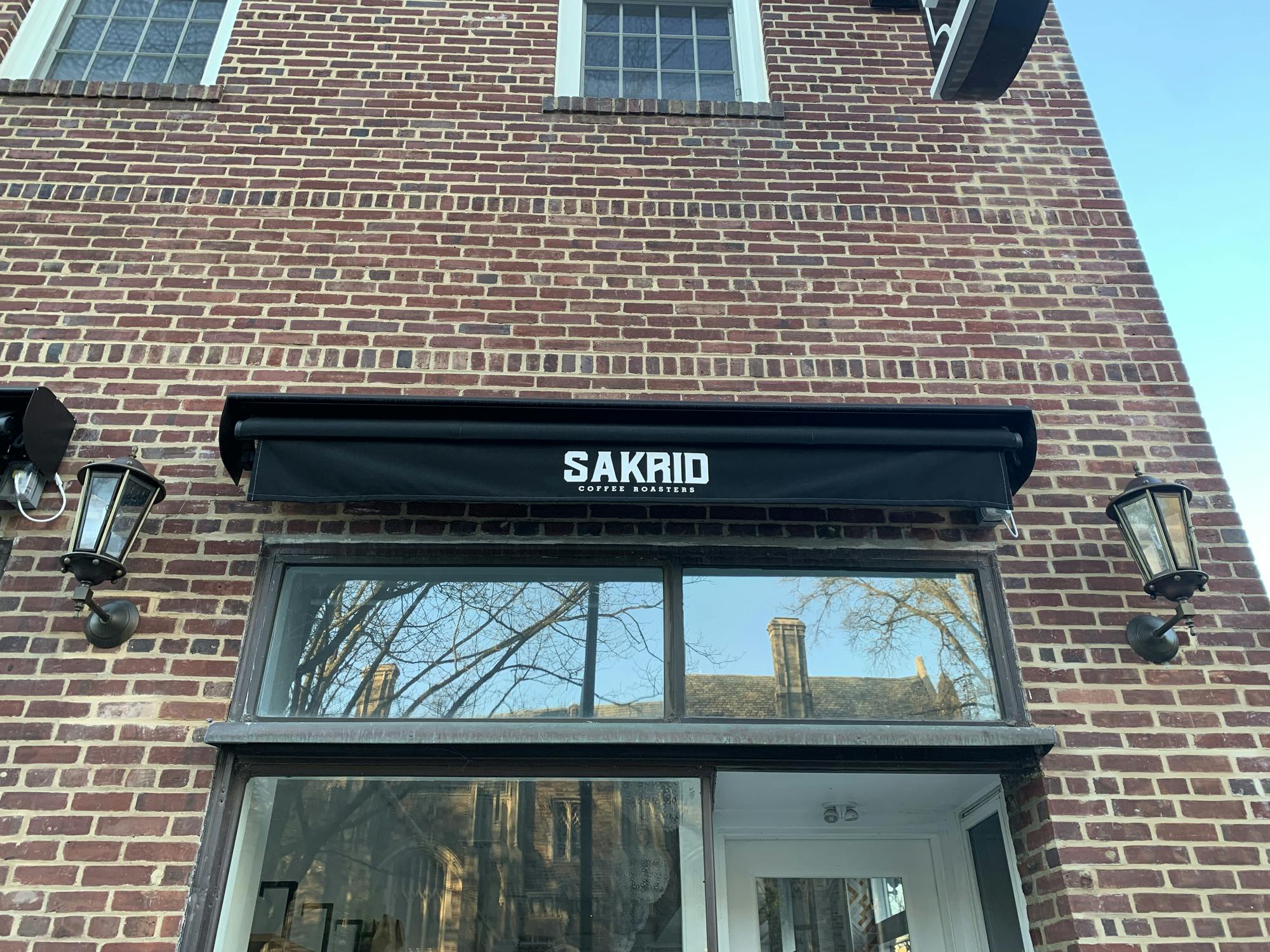 A brick building has a window with a draped curtain and the words “Sakrid Coffee Roasters” in fine print.   